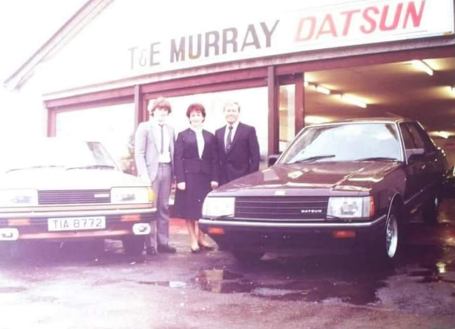 Tommy and Liz Murray standing outside their Datsun garage with their son Seamus beside a new Datsun car.